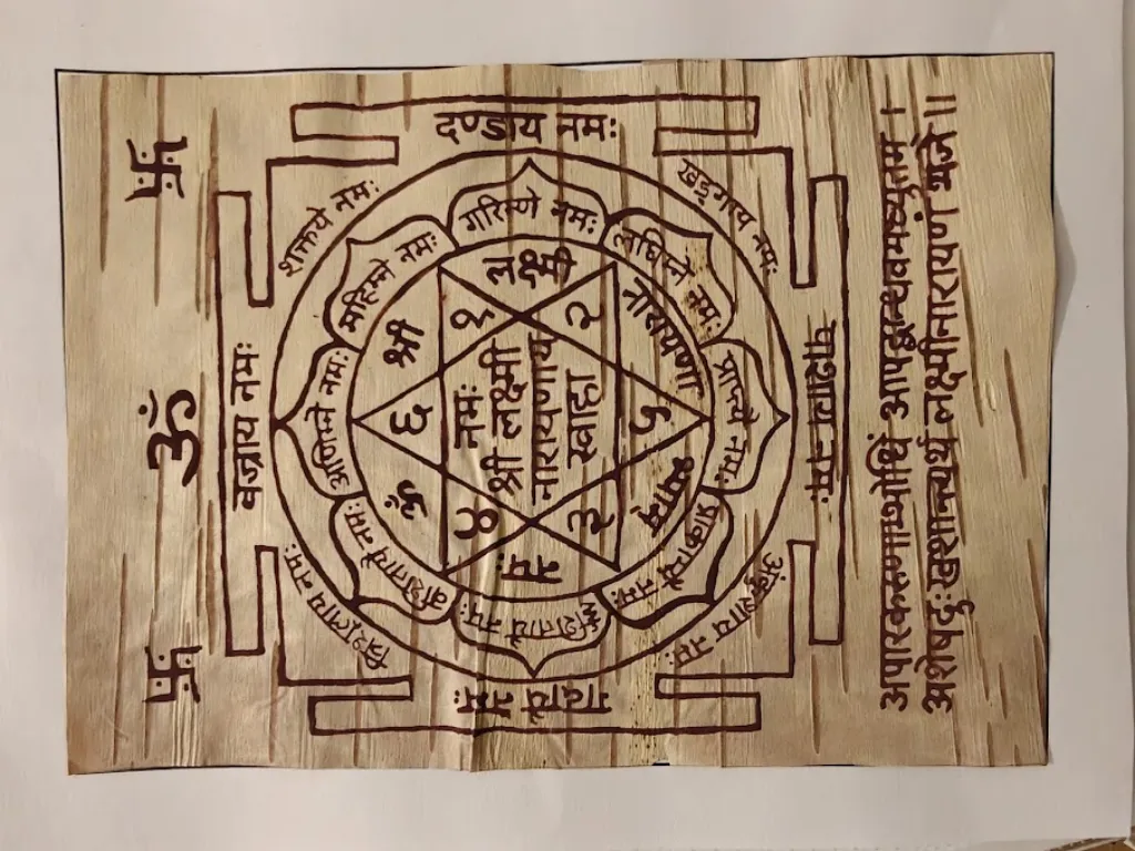 Vista cenital de un yantra terminado a mano en una lámina rectangular de bhojpatra del Himalaya. El diseño central presenta una estrella geométrica compleja dentro de círculos concéntricos y un marco cuadrado bhupura con cuatro puertas ceremoniales. Incluye caligrafía sánscrita trazada con tinta tradicional de ashtagandha de color granate, con el mantra central Om Namah Shri Lakshmi Narayanaya Swaha. A la izquierda aparecen símbolos sagrados como Om y esvástica; a la derecha, un verso vertical en sánscrito.