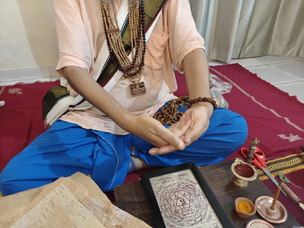 Vista desde arriba de Shri Damodar Dasji Maharaj sentado con un dhoti azul mientras realiza la prana-pratishtha. Sus manos forman un mudra específico de energización sobre un yantra dibujado a mano en bhojpatra del Himalaya. El yantra está enmarcado y colocado sobre una superficie de madera junto a elementos tradicionales, como akshat, haldi y una lámpara con tinta natural. Lleva varias malas sagradas y un colgante de Sita-Ram, representativos de la tradición Ramanandi.