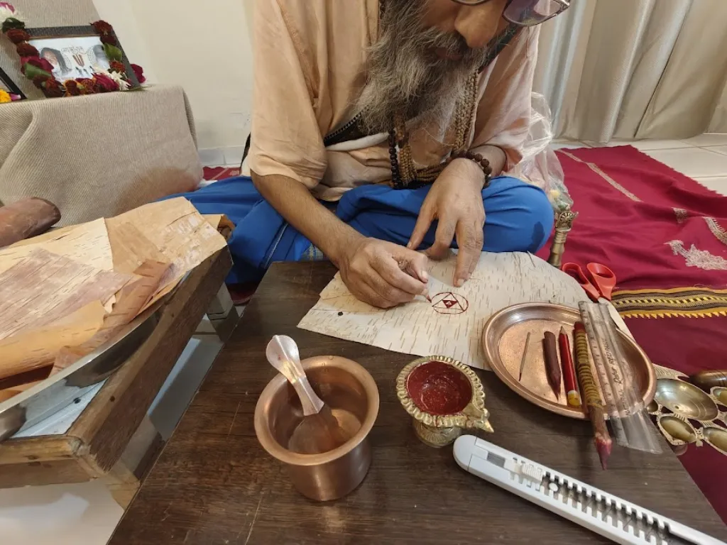 Vista cercana desde arriba de Shri Damodar Dasji Maharaj, un maestro de barba gris vestido con ropa espiritual tradicional de tonos azafrán y azul, dibujando cuidadosamente un yantra geométrico a mano. Está sentado en el suelo y utiliza un cálamo tradicional sobre auténtico bhojpatra del Himalaya (corteza de abedul). Frente a él se encuentran un pequeño recipiente de cobre con agua, una lámpara ritual con pasta roja, una regla y varios cálamos naturales. Al fondo hay una imagen sagrada enmarcada, decorada con una guirnalda de flores, sobre un altar.