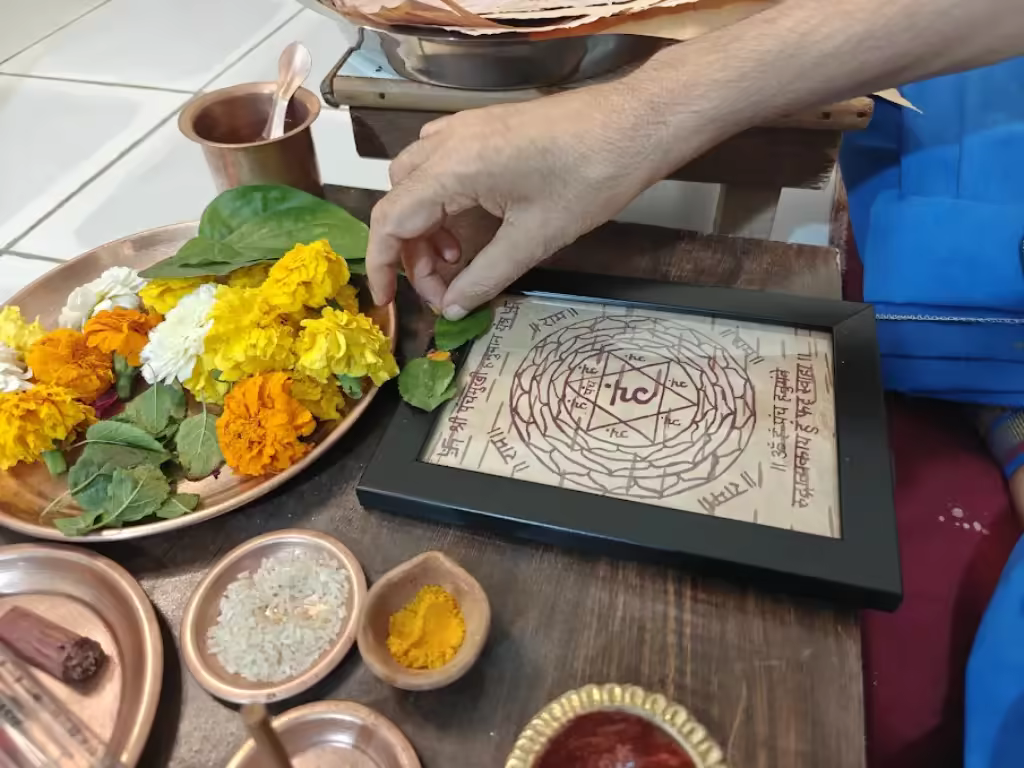 A close-up view of the Prana-Pratishtha process, showing the hand of Shri Damodar Dasji Maharaj placing a sacred Tulsi leaf (Tulsi Arpanam) onto a framed Panchmukhi Hanuman Yantra. The Yantra is hand-drawn on authentic Himalayan Bhojpatra with traditional natural inks. Surrounding the Yantra are consecrated elements of the Trust Stack: a plate of fresh marigold flowers, small containers of Akshat (consecrated rice), Haldi (turmeric), and a pomegranate twig stylus used for the sacred geometry.